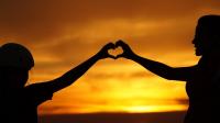 Teenager and a caregiver making a hand heart together in front of a sunset.