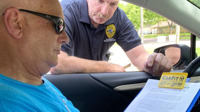 Older man fills out form while sitting inside car as younger man leans in window to discuss